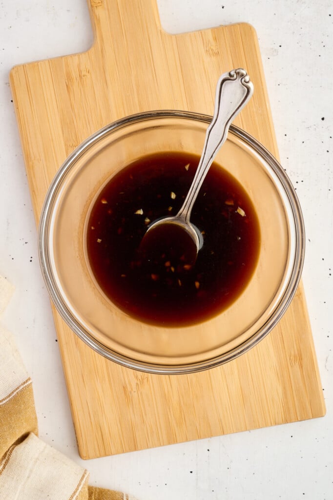 A glass bowl filled with dark sauce and minced garlic, perfect for marinating Healthy High Protein Grilled Teriyaki Chicken & Pineapple Skewers, with a metal spoon inside, sits on a wooden cutting board atop a white surface.