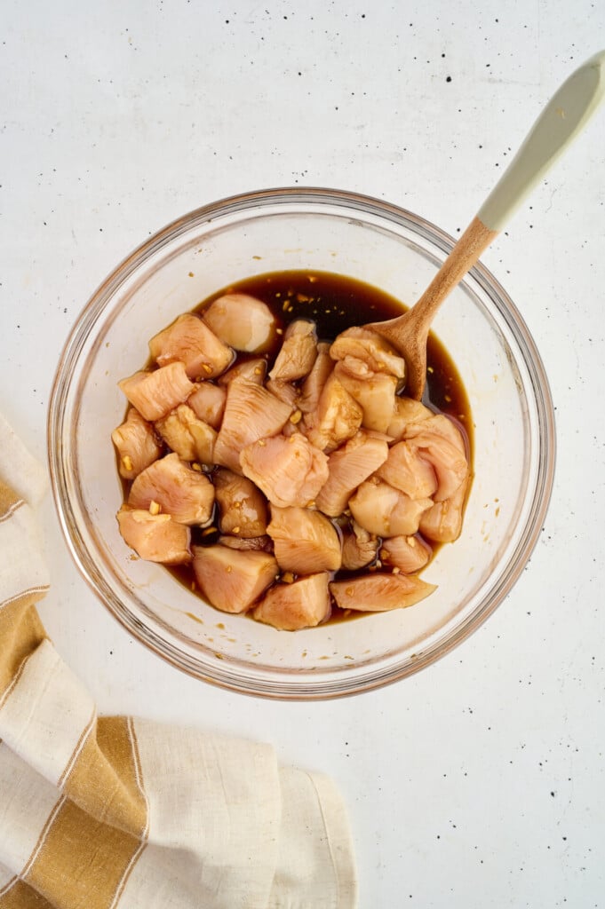 Raw chicken pieces, soon to become Healthy High Protein Grilled Teriyaki Chicken & Pineapple Skewers, marinate in a dark sauce in a glass bowl with a wooden spoon, placed on a white surface next to a beige and white striped kitchen towel.