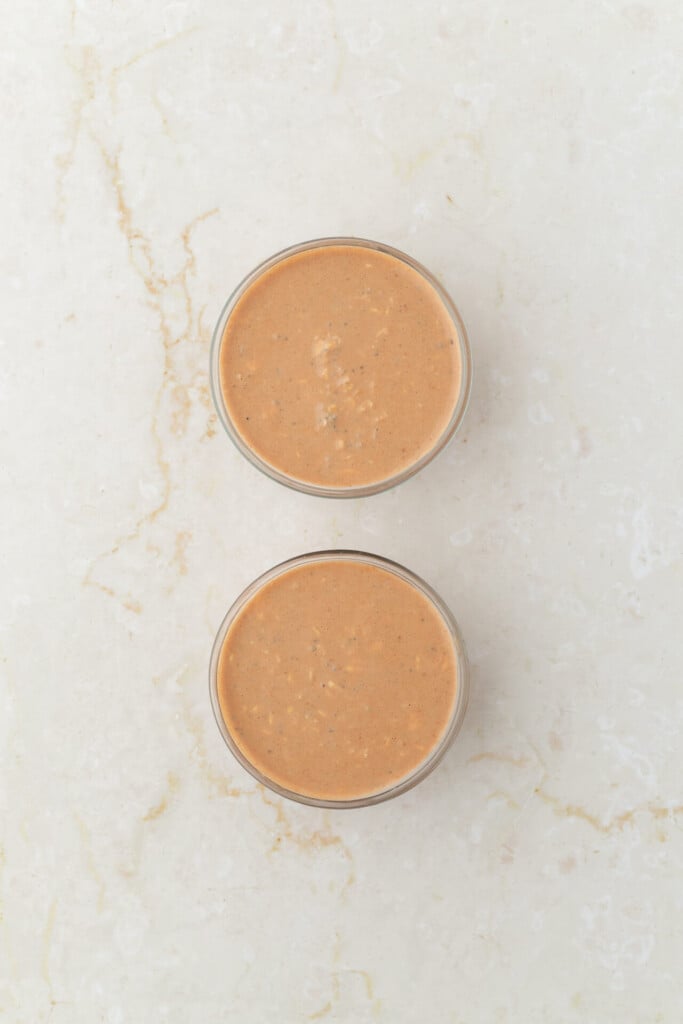Two glass cups filled with a creamy light brown smoothie reminiscent of Chocolate Peanut Butter Overnight Oats are placed on a light marble surface, viewed from above.