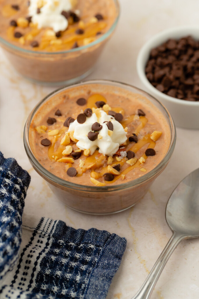 A glass bowl of creamy dessert, reminiscent of Chocolate Peanut Butter Overnight Oats, topped with whipped cream, chocolate chips, caramel drizzle, and chopped nuts, with a spoon, blue napkin, and a bowl of chocolate chips nearby.