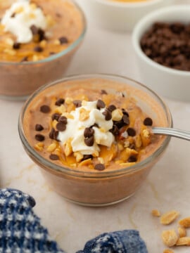 A glass bowl of Chocolate Peanut Butter Overnight Oats is topped with whipped cream, chocolate chips, chopped nuts, and a drizzle of peanut butter. A spoon rests in the bowl while other tempting toppings are visible in the background.