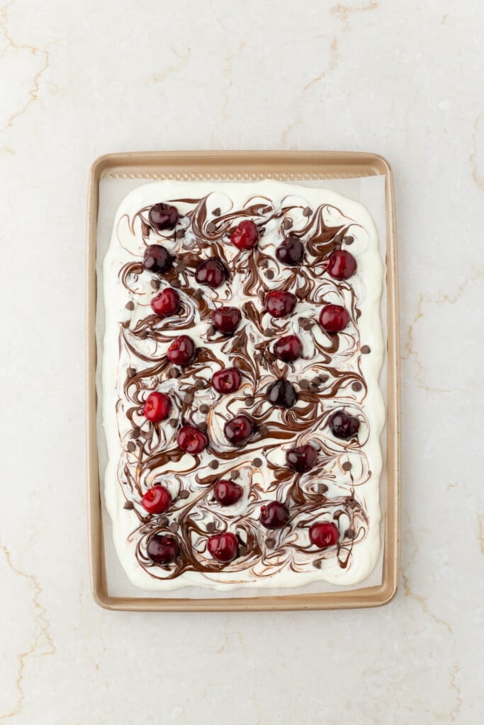 A rectangular baking tray of Frozen Chocolate Cherry Yogurt Bark, topped with creamy swirls of chocolate and scattered fresh cherries, set on a light marble surface.