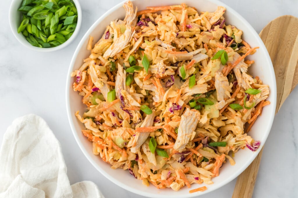 A bowl of shredded chicken coleslaw with carrots, cabbage, and green onions, served on a white surface with a wooden spoon and a small bowl of chopped green onions nearby.