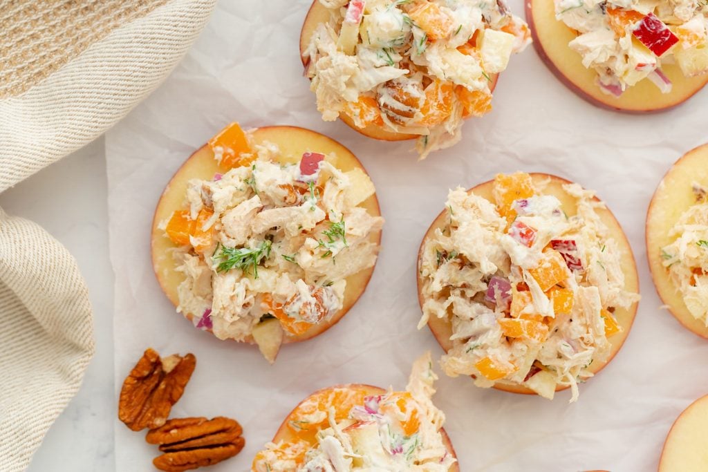 Apple slices topped with a creamy chicken salad containing diced vegetables and herbs, arranged on parchment paper, with a few pecan halves beside them. A textured beige cloth is visible in the corner.