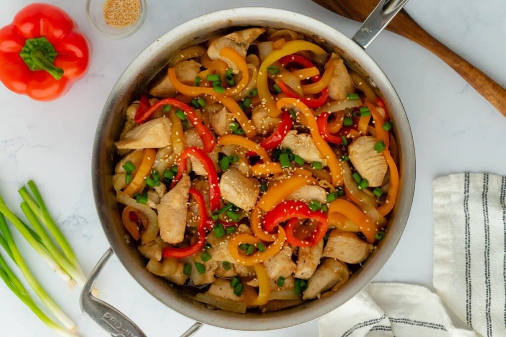 A skillet filled with stir-fried chicken, red and yellow bell peppers, topped with sesame seeds and chopped green onions. A wooden spoon, green onions, a red pepper, and a striped towel are nearby on a marble surface.