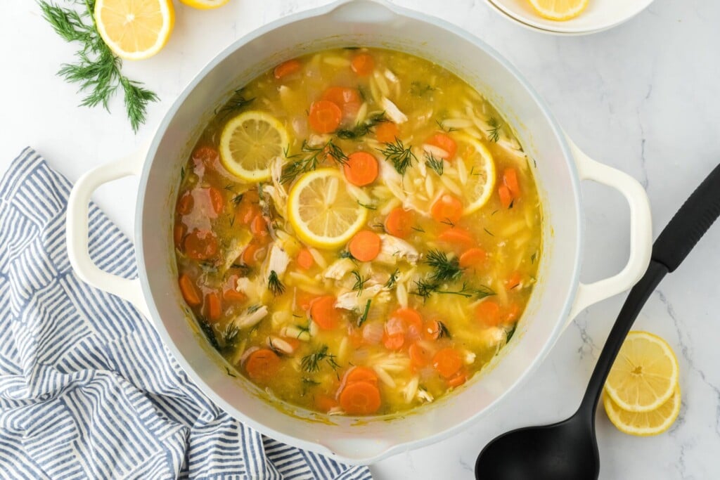 A pot of chicken orzo soup with sliced carrots, lemon rounds, and fresh dill, on a marble counter with lemon slices, herbs, a striped towel, and a black ladle nearby.