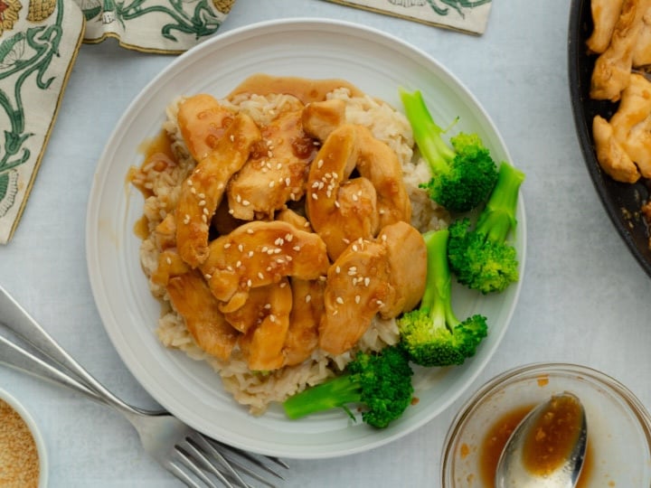 A plate of rice topped with teriyaki chicken strips, sesame seeds, and broccoli florets, with a fork and napkin nearby.