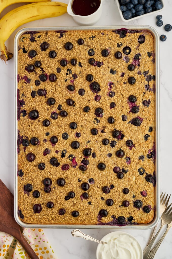 A large rectangular tray of Easy Sheet Pan Blueberry Oatmeal Pancakes topped with blueberries sits on a marble surface, surrounded by bananas, a bowl of blueberries, syrup, and whipped cream.