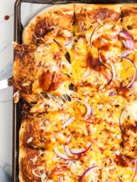 A baked sheet pan pizza topped with melted cheese, sliced red onions, and barbecue sauce sits on a pan. A metal spatula lifts a cheesy slice from the corner.