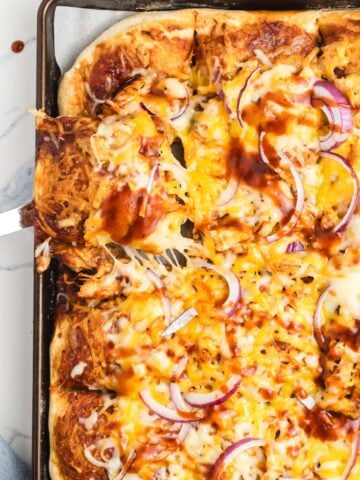 A baked sheet pan pizza topped with melted cheese, sliced red onions, and barbecue sauce sits on a pan. A metal spatula lifts a cheesy slice from the corner.