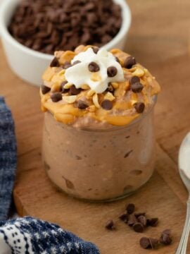 A jar of Chocolate Peanut Butter Overnight Oats topped with whipped cream and chocolate chips sits on a wooden board next to a spoon, a blue patterned napkin, and small bowls of oats, chocolate chips, and peanut butter.