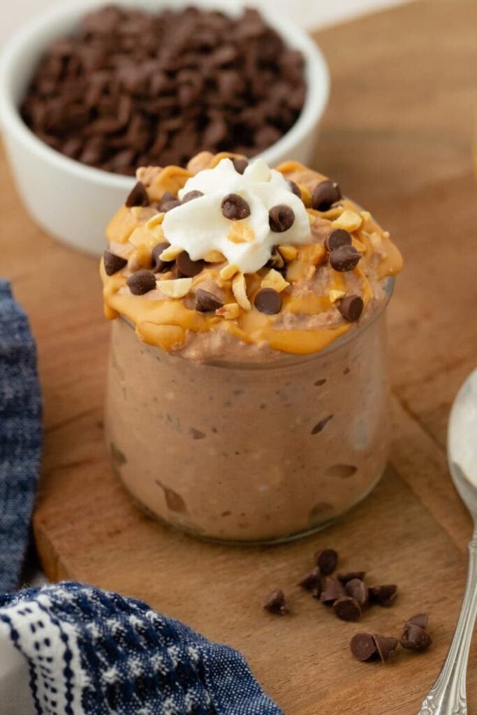 A jar of Chocolate Peanut Butter Overnight Oats topped with whipped cream and chocolate chips sits on a wooden board next to a spoon, a blue patterned napkin, and small bowls of oats, chocolate chips, and peanut butter.