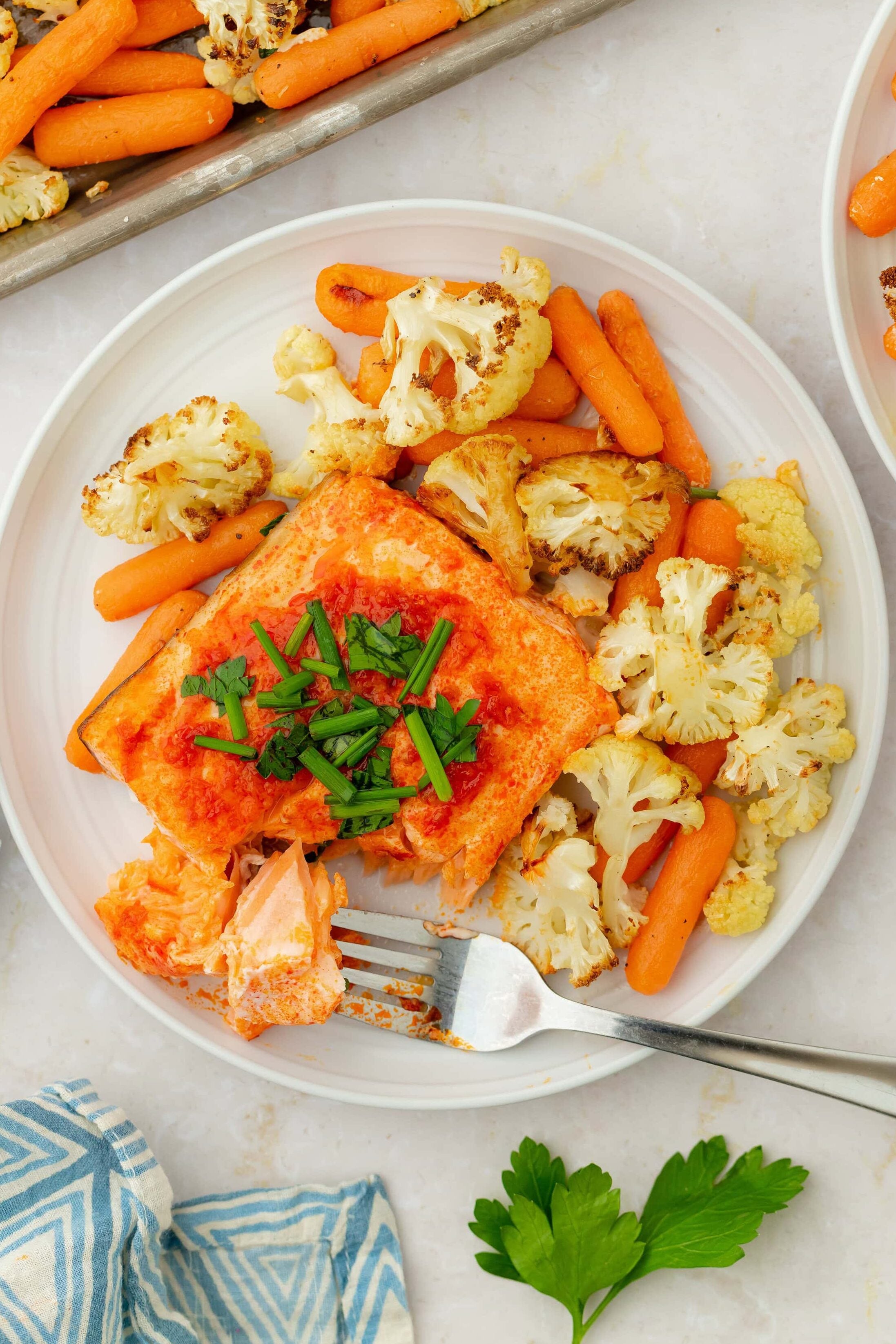 A plate of Sheet Pan Harissa Salmon with Veggies features baked salmon topped with chopped chives, roasted cauliflower, and baby carrots. A fork holds a piece of salmon, while a blue patterned napkin and parsley sit nearby.