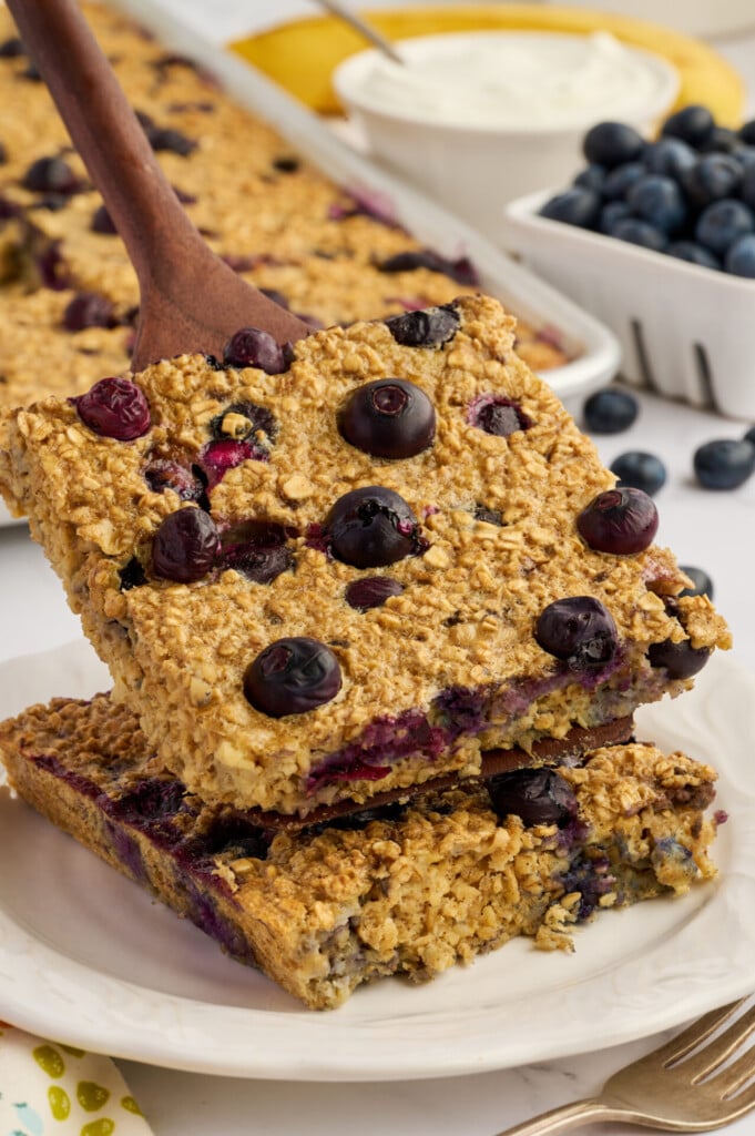 Two oat and blueberry baked squares are stacked on a plate, with a wooden spatula lifting the top square. Reminiscent of Easy Sheet Pan Blueberry Oatmeal Pancakes, this delicious treat is surrounded by fresh blueberries and a dish of yogurt in the background.