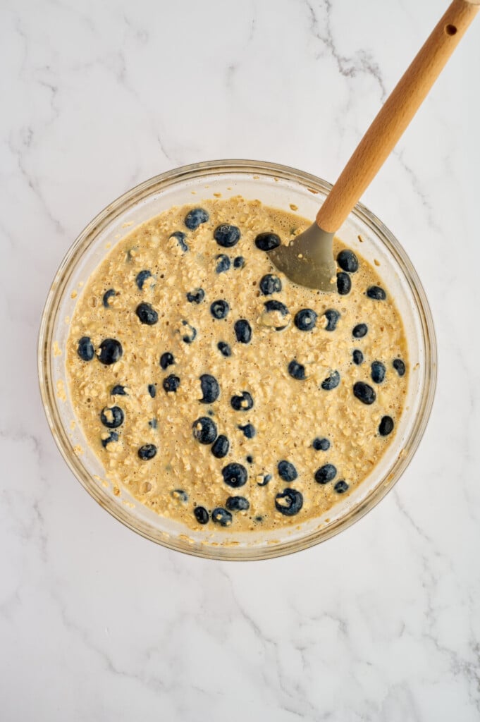 A glass bowl filled with blueberry muffin batter for Easy Sheet Pan Blueberry Oatmeal Pancakes and a spatula on a white marble surface.