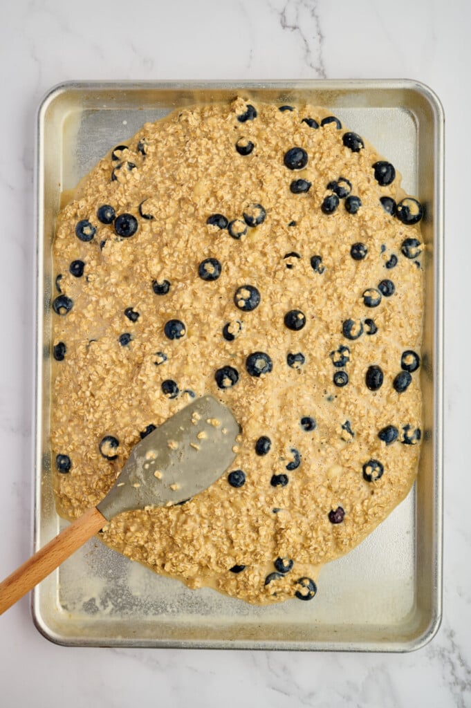 A baking sheet with Easy Sheet Pan Blueberry Oatmeal Pancakes batter spread out, a spatula resting on the sheet, all on a white marble surface.