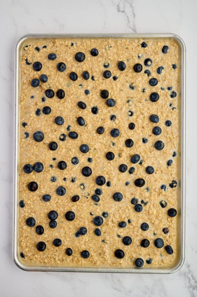 A baking sheet filled with unbaked Easy Sheet Pan Blueberry Oatmeal Pancakes batter, topped with blueberries, sitting on a white marble surface.