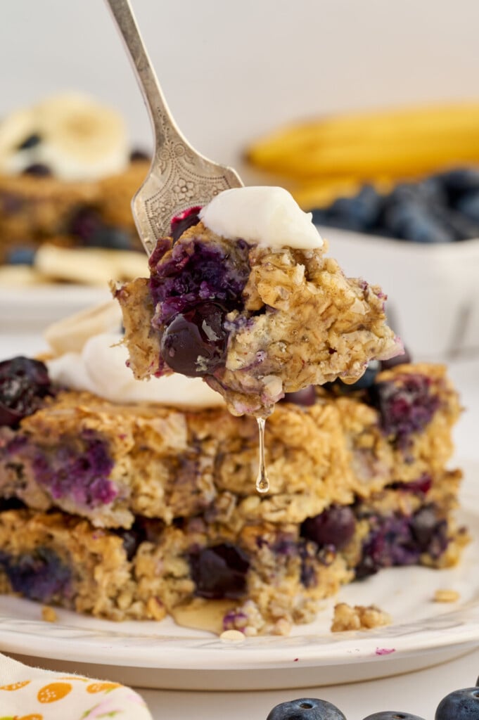 A fork holds a bite of Easy Sheet Pan Blueberry Oatmeal Pancakes with blueberries and a dollop of cream above a stack of similar oatmeal squares, with bananas and blueberries in the background.