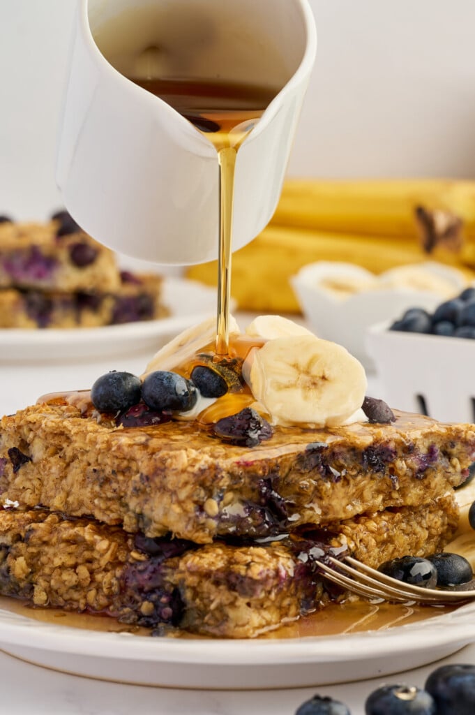 Syrup being poured over stacked Easy Sheet Pan Blueberry Oatmeal Pancakes, topped with fresh blueberries and banana slices, with bananas and blueberries in the background.