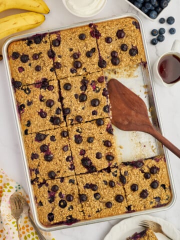 A baking pan of Easy Sheet Pan Blueberry Oatmeal Pancakes, partially served, sits on a table with a wooden spatula. Surrounding the pan are bananas, blueberries, syrup, a bowl of yogurt, and a floral-patterned napkin with forks.