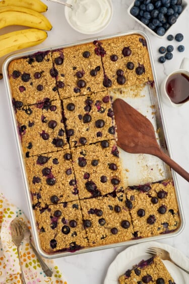 A baking pan of Easy Sheet Pan Blueberry Oatmeal Pancakes, partially served, sits on a table with a wooden spatula. Surrounding the pan are bananas, blueberries, syrup, a bowl of yogurt, and a floral-patterned napkin with forks.