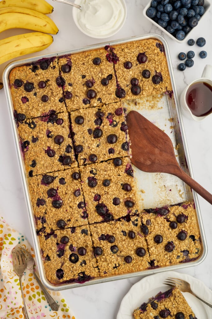 A baking pan of Easy Sheet Pan Blueberry Oatmeal Pancakes, partially served, sits on a table with a wooden spatula. Surrounding the pan are bananas, blueberries, syrup, a bowl of yogurt, and a floral-patterned napkin with forks.