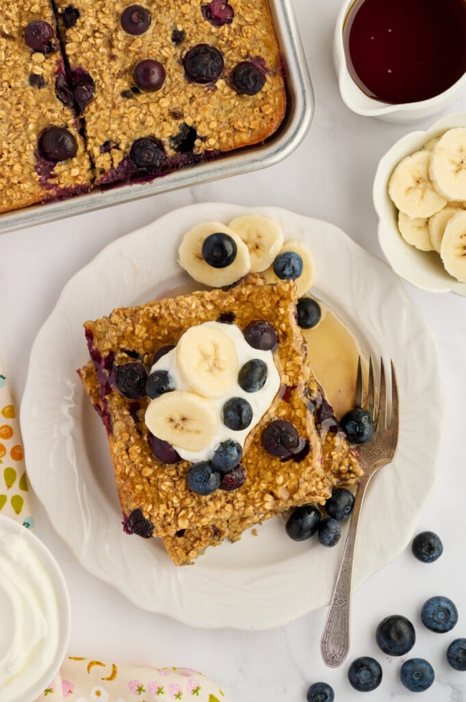 A slice of Easy Sheet Pan Blueberry Oatmeal Pancakes topped with banana slices and blueberries sits on a white plate with a fork. Nearby are a napkin, a bowl of banana slices, a bowl of syrup, and a dish of whipped cream.