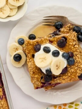 A slice of Easy Sheet Pan Blueberry Oatmeal Pancakes topped with banana slices and blueberries sits on a white plate with a fork. Nearby are a napkin, a bowl of banana slices, a bowl of syrup, and a dish of whipped cream.