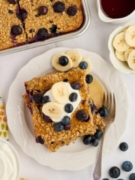 A slice of Easy Sheet Pan Blueberry Oatmeal Pancakes topped with banana slices and blueberries sits on a white plate with a fork. Nearby are a napkin, a bowl of banana slices, a bowl of syrup, and a dish of whipped cream.