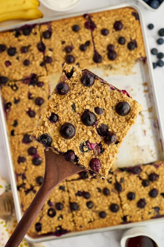 A wooden spatula holds a square of Easy Sheet Pan Blueberry Oatmeal Pancakes, filled with juicy blueberries, above a tray of similar blueberry oatmeal squares.