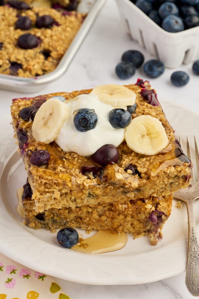 Two thick slices of Easy Sheet Pan Blueberry Oatmeal Pancakes with blueberries and bananas are stacked on a plate, topped with yogurt, banana slices, blueberries, and a drizzle of honey. A fork and a baking dish are nearby.