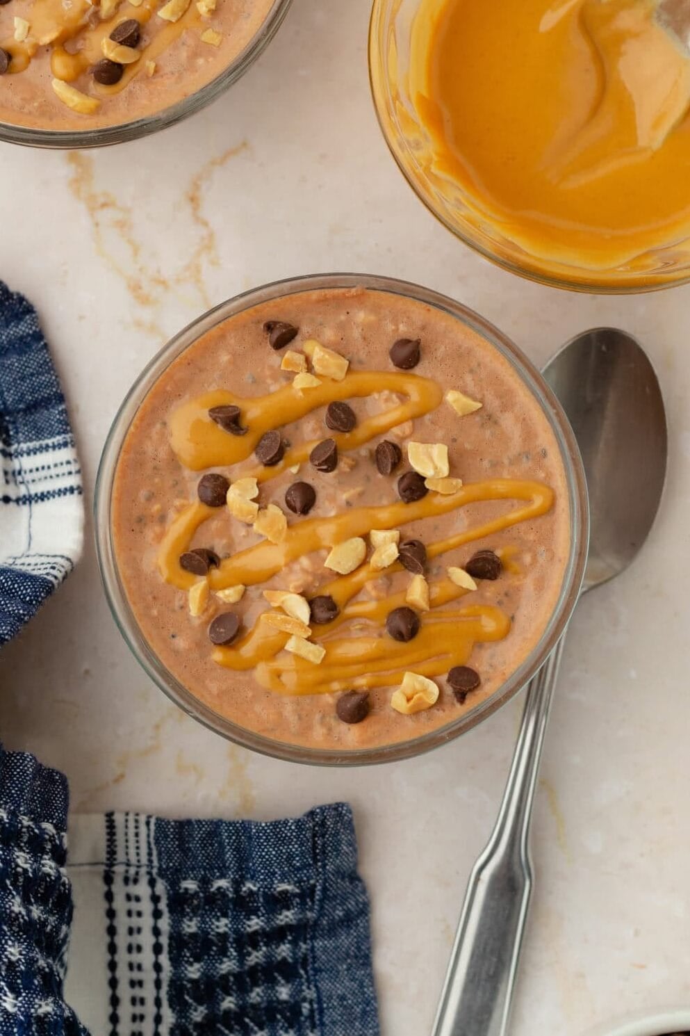 A bowl of chocolate pudding topped with chopped nuts, chocolate chips, and caramel sauce sits next to a spoon, a blue checkered cloth, a bowl of caramel sauce, and a small bowl of coffee beans—an indulgent scene that’s reminiscent of the decadent flavors found in Chocolate Peanut Butter Overnight Oats.