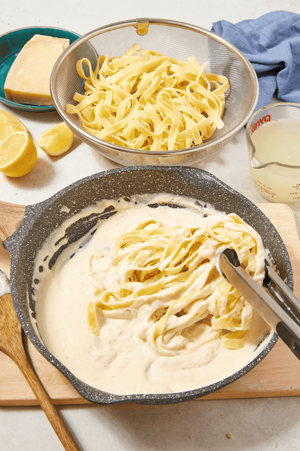 A skillet of high-protein Cottage Cheese Fettuccine Alfredo sauce with fettuccine being tossed in, next to a strainer of cooked pasta, Parmesan cheese, lemon wedges, and a measuring cup on a wooden board.