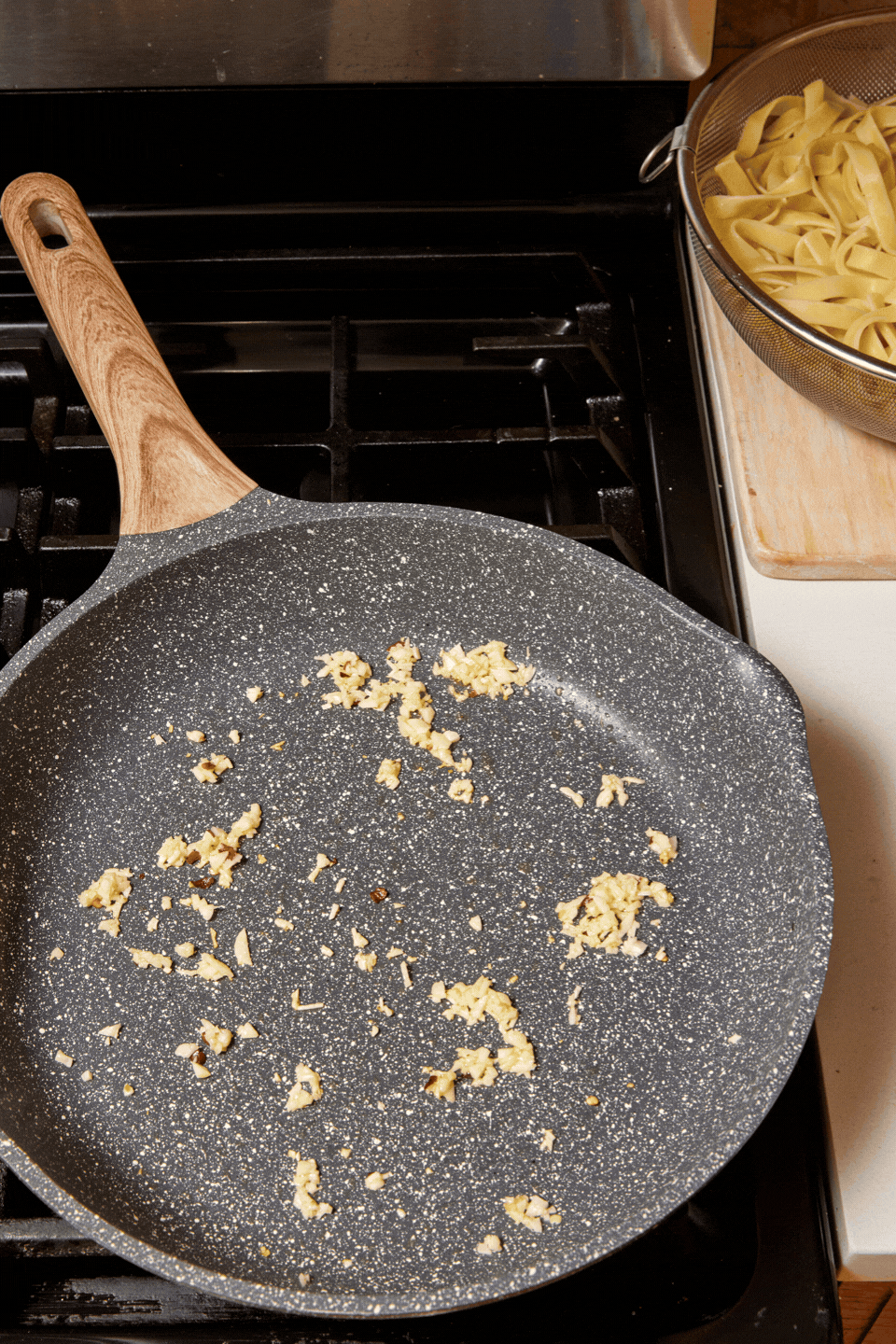 A frying pan with bits of minced garlic sits on a stovetop. Next to it, a colander with cooked pasta rests on the counter, ready for high-protein Cottage Cheese Fettuccine Alfredo.