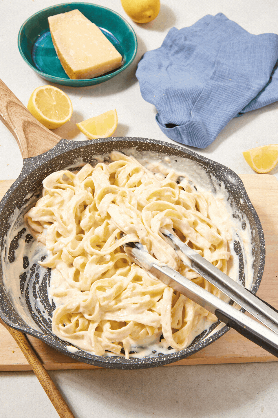 A pan of creamy high-protein Cottage Cheese Fettuccine Alfredo with metal tongs, surrounded by lemon wedges, a blue napkin, and a wedge of Parmesan cheese on a small green plate.