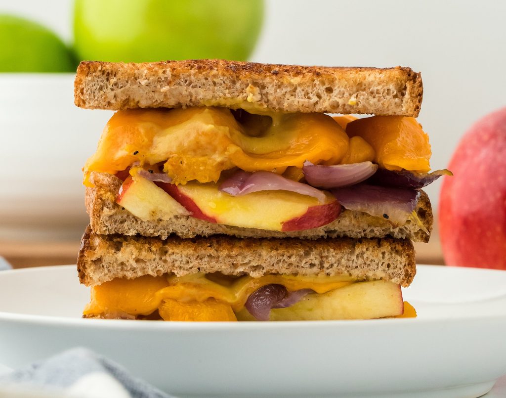 A close-up of a grilled cheese sandwich with melted cheddar, red onions, and apple slices on whole wheat bread—an ideal choice for healthy fall recipes—stacked on a white plate. Green apples and a peach are visible in the blurred background.