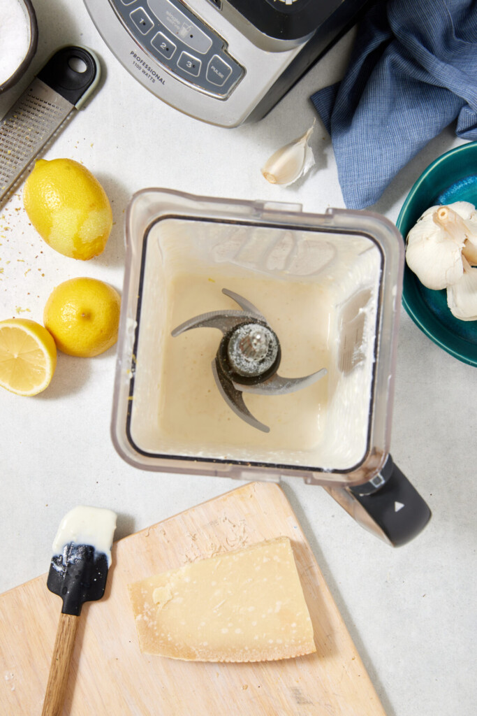 A blender with creamy high-protein Cottage Cheese Fettuccine Alfredo sauce sits on a counter near lemons, garlic bulbs, a blue towel, a black spatula, a wooden board, and Parmesan cheese.