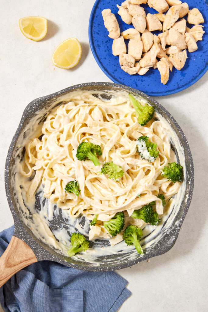 A skillet of high-protein Cottage Cheese Fettuccine Alfredo with broccoli sits beside a blue plate with pieces of cooked chicken and two lemon wedges on a light surface with a blue cloth.