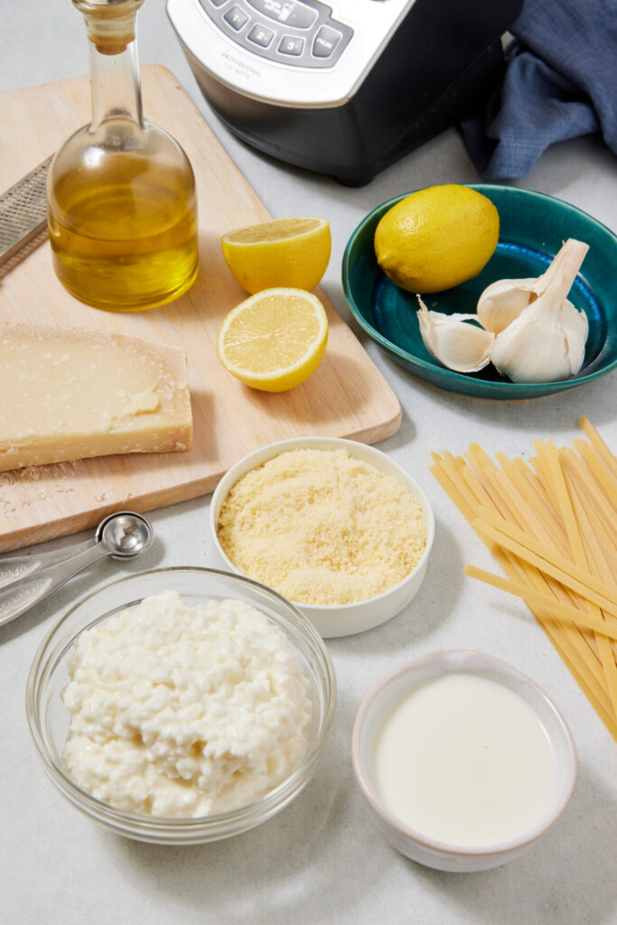 A cutting board holds olive oil, a wedge of cheese, lemon half, grated cheese, garlic, dry pasta, milk, and a bowl of ricotta—perfect for preparing high-protein Cottage Cheese Fettuccine Alfredo.
