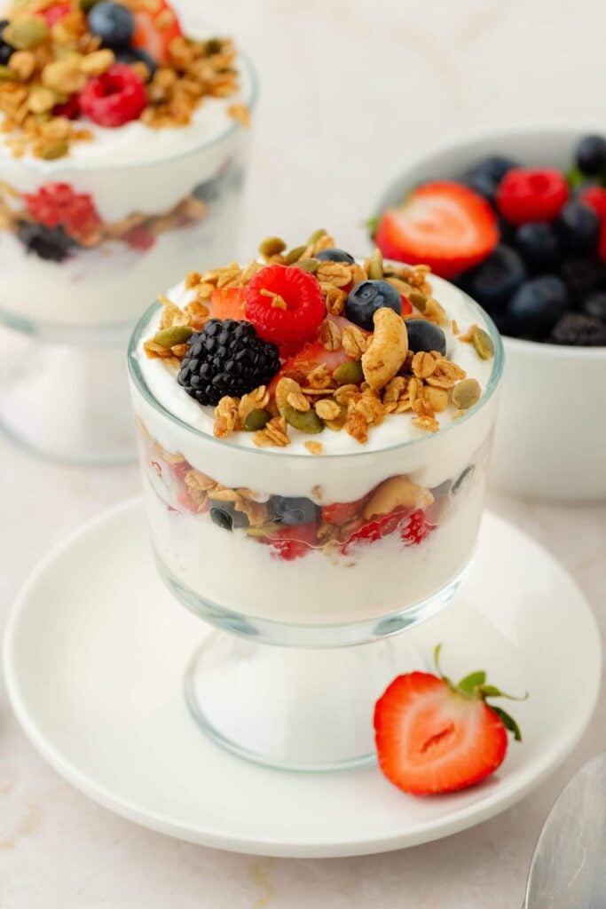 A glass cup filled with layers of Greek Yogurt Parfait, granola, and fresh berries sits on a white plate with a sliced strawberry. In the background, another parfait and a bowl of mixed berries are visible, alongside a spoon and napkin.