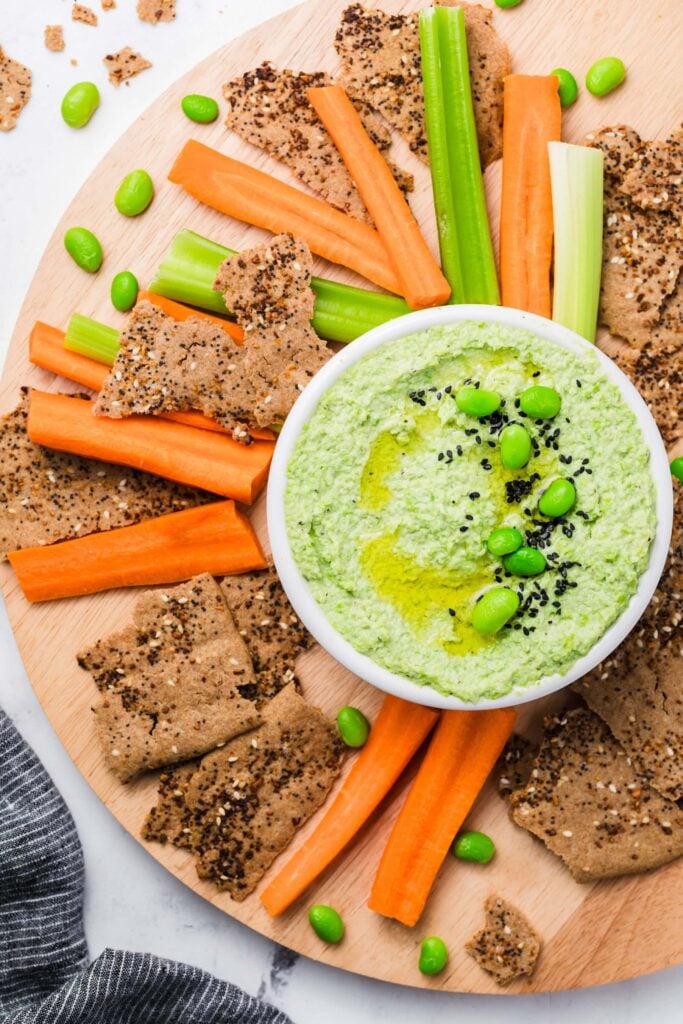 A round wooden platter features a bowl of creamy Edamame Hummus topped with edamame and black sesame seeds, surrounded by carrot and celery sticks, crackers, and scattered edamame beans.