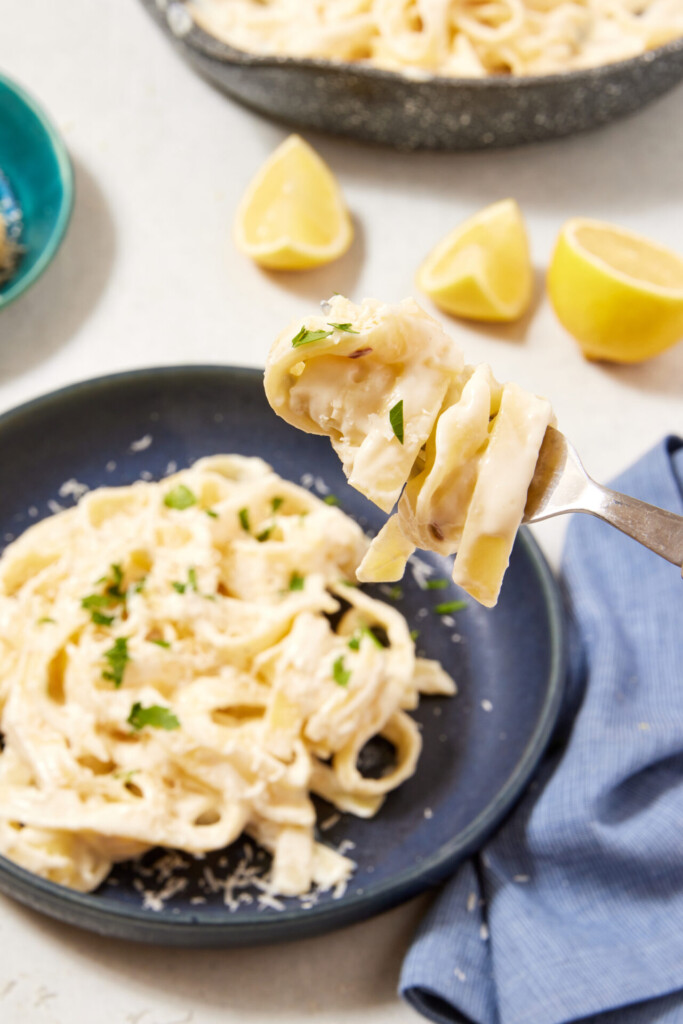 A fork holding creamy high-protein Cottage Cheese Fettuccine Alfredo hovers over a dark plate filled with pasta, garnished with parsley, with lemon wedges and a dish towel nearby.