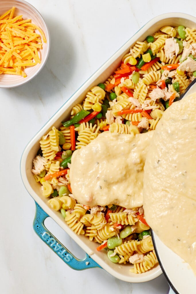 A casserole dish filled with rotini pasta, mixed vegetables, and chicken is topped with creamy sauce. A bowl of shredded cheese sits nearby on a white countertop.