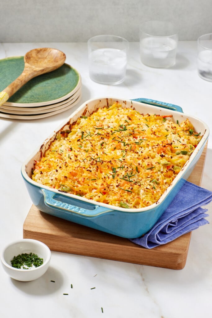 A baked casserole topped with golden, crispy cheese and herbs sits in a blue dish on a wooden board. Plates, a spoon, water glasses, and a small bowl of chopped herbs are in the background.