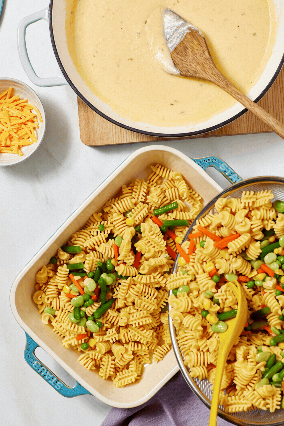 A casserole dish filled with pasta and mixed vegetables next to a bowl of shredded cheese and a pot of creamy sauce being stirred with a wooden spoon.