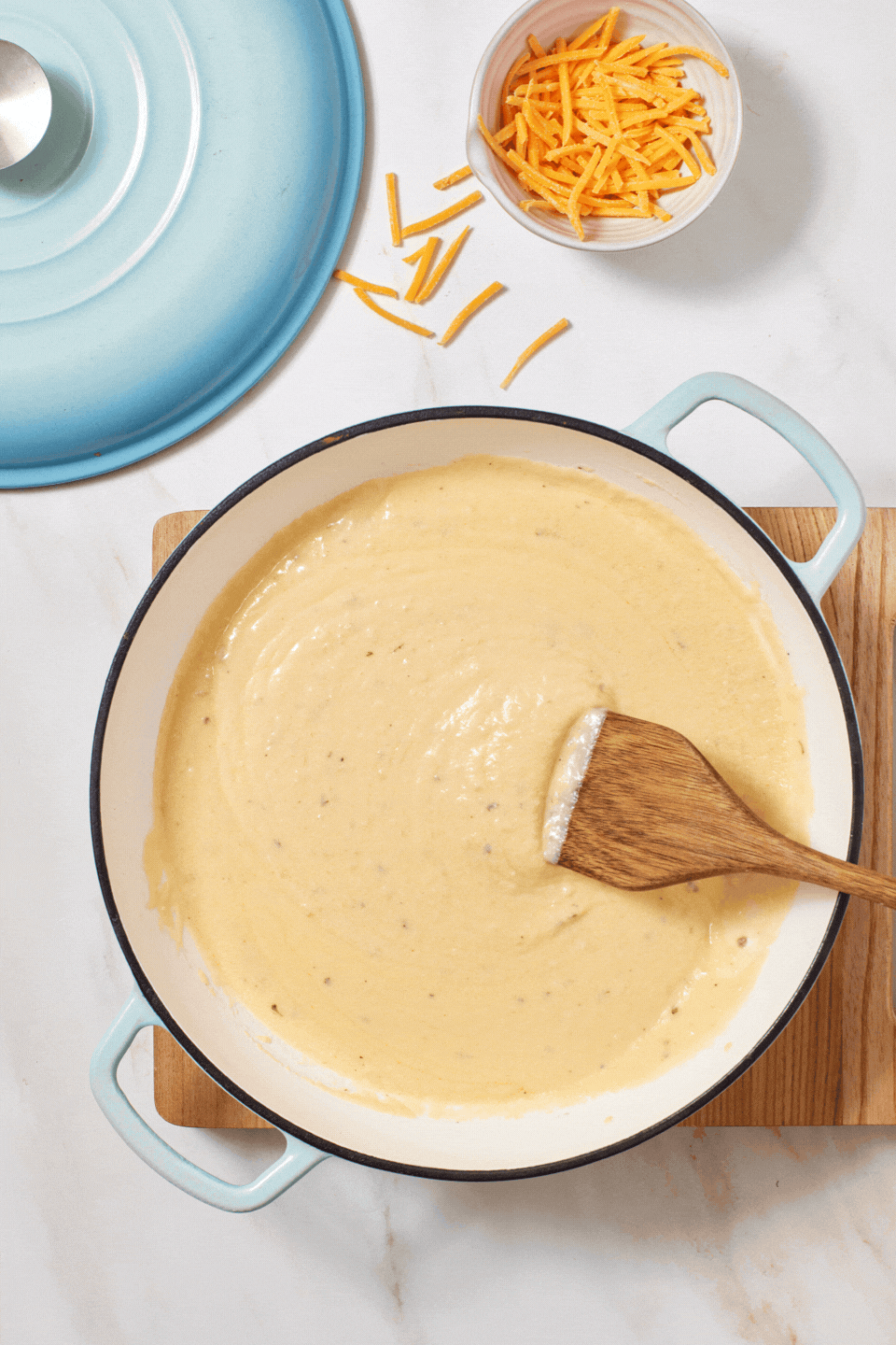 A creamy cheese sauce is being stirred in a white pot with a wooden spatula. A small bowl of shredded cheddar cheese and a blue lid are nearby on a marble surface.