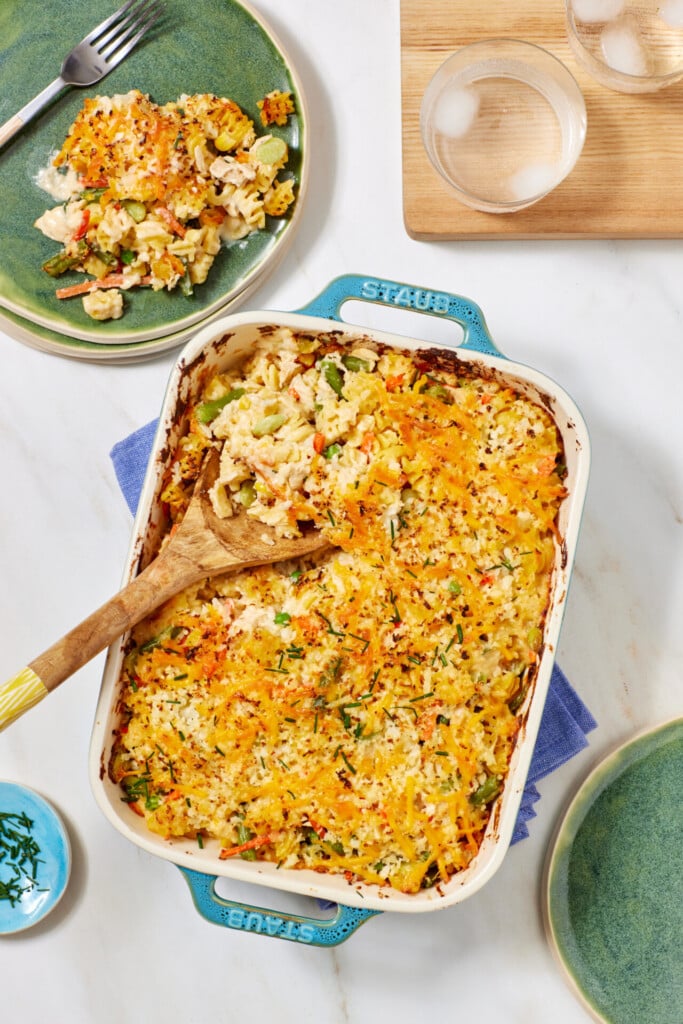 Casserole dish filled with cheesy baked vegetable casserole, partially served with a wooden spoon. A portion is plated on a green plate beside two empty plates and two glasses of water with ice.