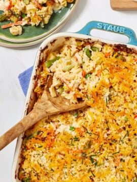 Casserole dish filled with cheesy baked vegetable casserole, partially served with a wooden spoon. A portion is plated on a green plate beside two empty plates and two glasses of water with ice.