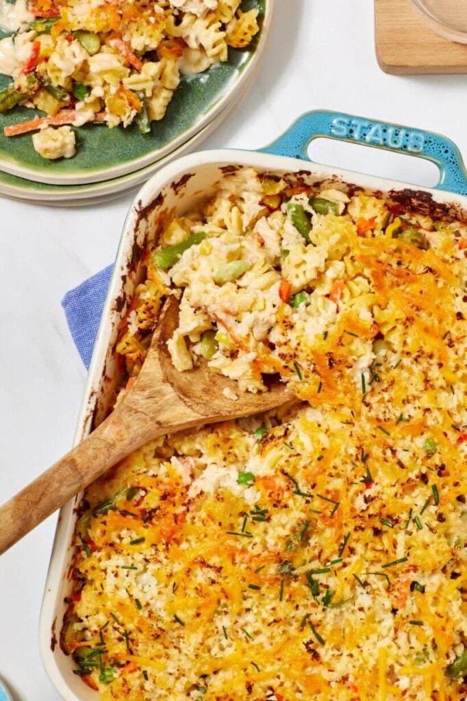 Casserole dish filled with cheesy baked vegetable casserole, partially served with a wooden spoon. A portion is plated on a green plate beside two empty plates and two glasses of water with ice.