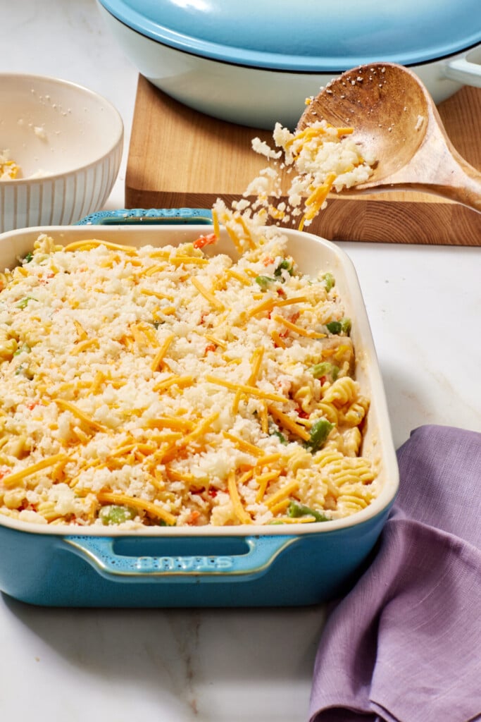 A blue baking dish filled with a pasta casserole topped with shredded cheese. A wooden spoon sprinkles more cheese on top. A purple napkin and another dish are nearby.